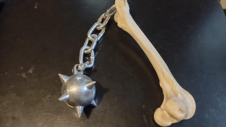May include: A medieval-style mace with a spiked metal ball attached to a chain, next to a light-colored bone. The mace and bone are set against a dark background. The mace is a weapon.