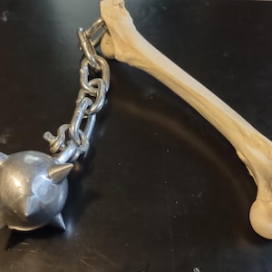 May include: A medieval-style weapon featuring a bone handle and a spiked metal ball connected by a chain. The bone is light brown, and the ball is silver. The chain is made of silver links. The weapon is likely a mace.