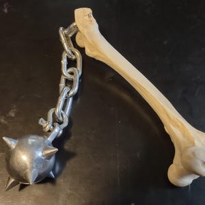 May include: A medieval-style flail weapon. The weapon features a wooden handle, a metal chain, and a spiked metal ball. The handle is a light tan color, and the metal components are silver.