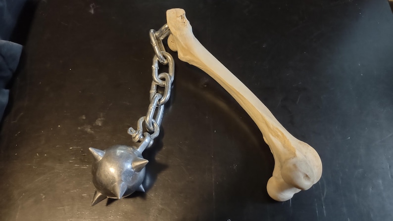 May include: A medieval-style flail weapon. The weapon features a wooden handle, a metal chain, and a spiked metal ball. The handle is a light tan color, and the metal components are silver.