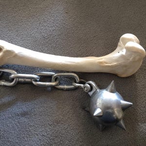 May include: A medieval-style flail weapon. The flail features a bone handle connected to a chain and a spiked metal ball. The bone is off-white, the chain is silver, and the ball is metallic.