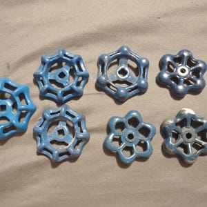 Puede incluir: Un conjunto de siete manijas de válvulas de agua azules, cada una con un diseño único en forma de estrella. Las manijas son de metal y varían ligeramente en tamaño y desgaste, lo que sugiere que son vintage o recicladas.