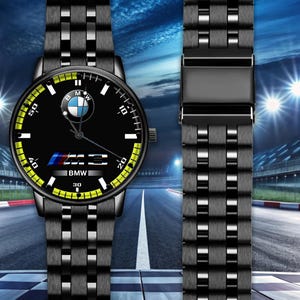 May include: Black wristwatch with a black dial featuring the BMW logo, M3 emblem, and BMW text. The watch has a yellow and white minute track and a black metal band. The background shows a racetrack.
