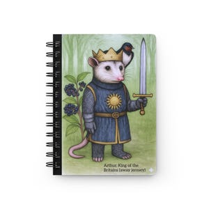 May include: Spiral-bound notebook featuring an illustration of an opossum dressed as a king, holding a sword, with a bird on its head. The opossum wears a crown and a blue tunic with a sun emblem. Text reads "Arthur, King of the Britains (away jerese)".