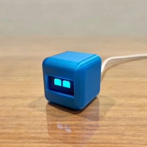 May include: A small, bright blue cube-shaped electronic device with a smooth matte finish. The front face features a dark rectangular screen displaying two glowing cyan square icons. A thin white power cable extends from the back of the device.