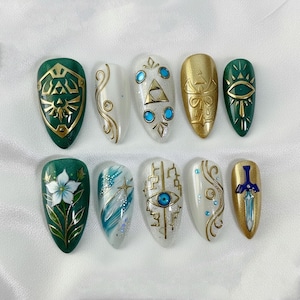 May include: A set of ten artificial nails with various designs. The nails feature gold and green shield, eye, and sword designs, along with floral and abstract patterns. Some nails have a white base with gold accents and blue gems.