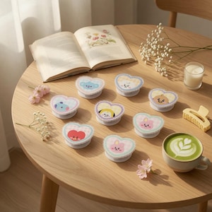 May include: A collection of heart-shaped phone grips with cartoon character designs in various colors. The grips are arranged on a wooden table with a book, flowers, and a cup of matcha latte.