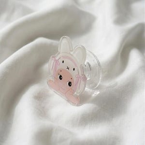 May include: A clear baby pacifier with a cartoon illustration of a baby wearing a bunny hat. The illustration features pink accents and a cross on the hat. The pacifier is resting on a white, textured surface.