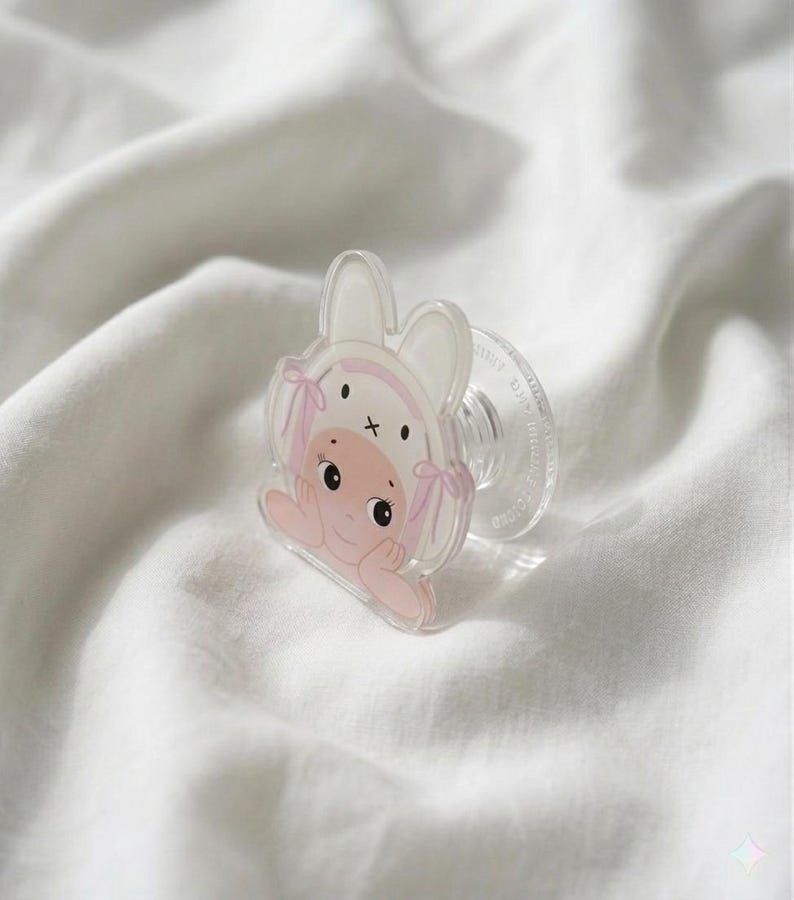 May include: A clear baby pacifier with a cartoon illustration of a baby wearing a bunny hat. The illustration features pink accents and a cross on the hat. The pacifier is resting on a white, textured surface.