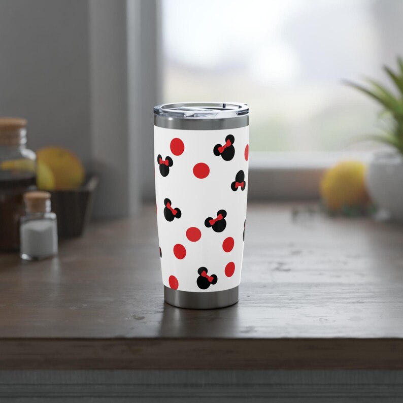 May include: A white travel tumbler with a silver lid, adorned with a pattern of red polka dots and black Minnie Mouse silhouettes with red bows. The tumbler is stainless steel and appears to be insulated.