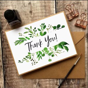 May include: A white card with the words "Thank You!" in black script, surrounded by green watercolor leaves. The card is on a brown envelope, with a paintbrush, ink bottle, and copper binder clips nearby.
