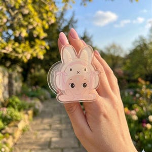 May include: A clear, pink bunny-themed phone grip. The grip features a cartoon bunny design with a baby bunny on top. The background is a blurred outdoor scene with greenery and a blue sky.