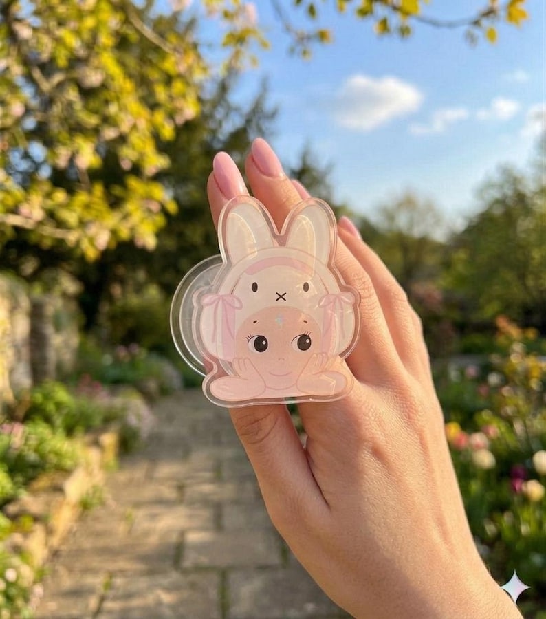 May include: A clear, pink bunny-themed phone grip. The grip features a cartoon bunny design with a baby bunny on top. The background is a blurred outdoor scene with greenery and a blue sky.