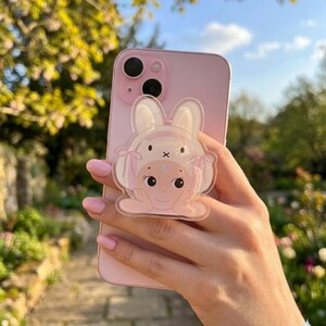 May include: A pink iPhone with a clear bunny-shaped phone grip. The grip features a cartoon bunny face with a bow and a star on its forehead. The phone is held in a hand with pink nail polish, with a blurred background of greenery.