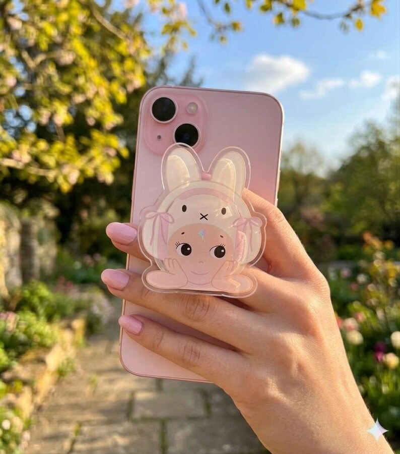 May include: A pink iPhone with a clear bunny-shaped phone grip. The grip features a cartoon bunny face with a bow and a star on its forehead. The phone is held in a hand with pink nail polish, with a blurred background of greenery.
