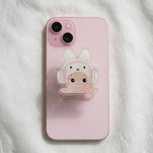 May include: A light pink smartphone with a cartoon bunny-themed phone grip. The grip features a smiling baby face wearing a bunny hat with pink bows. The phone has a dual-lens camera and is set against a white fabric background.