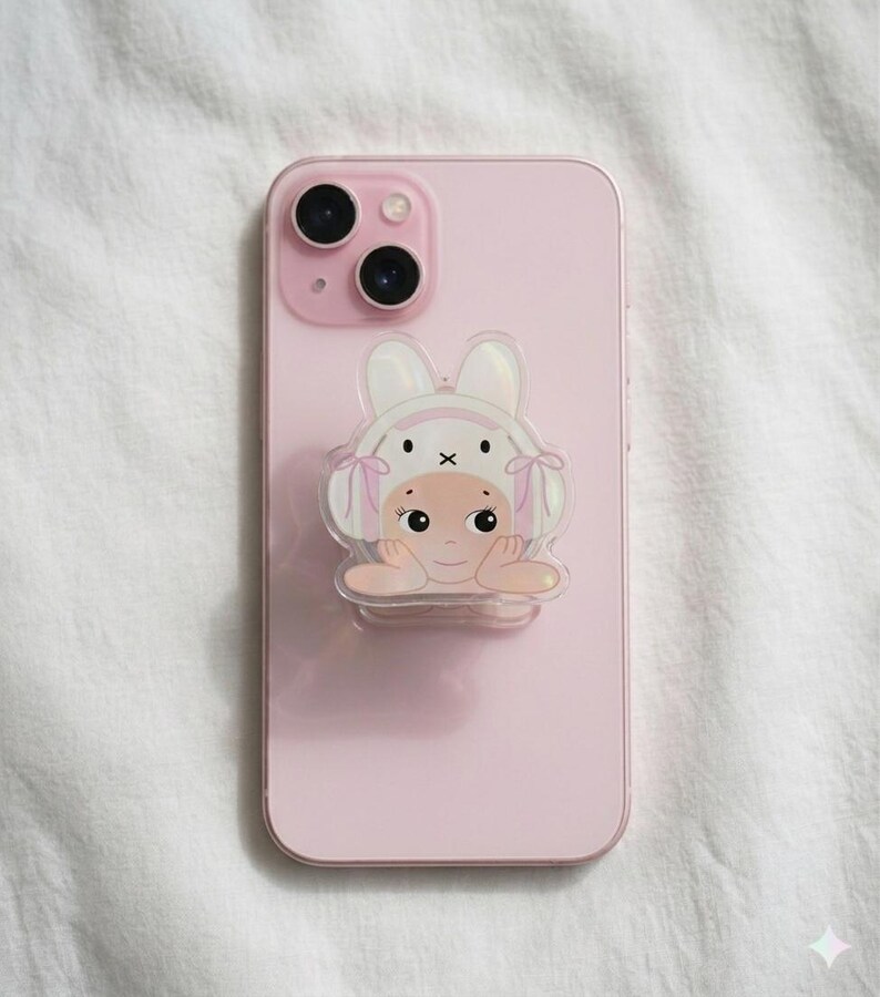 May include: A light pink smartphone with a cartoon bunny-themed phone grip. The grip features a smiling baby face wearing a bunny hat with pink bows. The phone has a dual-lens camera and is set against a white fabric background.
