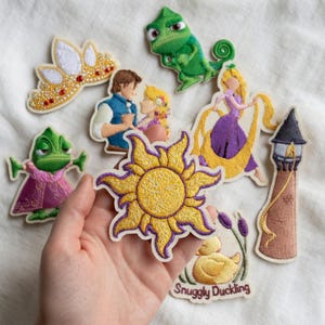 May include: Embroidered patches featuring characters and elements from a fairy tale. The collection includes a yellow sun, a tower, a crown, a duck, and characters in various poses. The patches are in shades of yellow, purple, green, and brown.