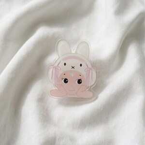 May include: A clear acrylic pin featuring a cartoon character wearing a bunny hat with pink accents. The character has a light pink complexion, blue star-shaped highlights, and is resting their chin on their hands. The pin is set against a white, textured background.