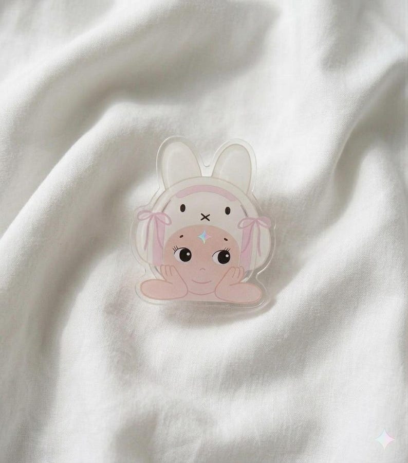 May include: A clear acrylic pin featuring a cartoon character wearing a bunny hat with pink accents. The character has a light pink complexion, blue star-shaped highlights, and is resting their chin on their hands. The pin is set against a white, textured background.