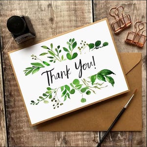 May include: A white card with the words "Thank You!" in black script, surrounded by green watercolor leaves. Includes a brown envelope, ink bottle, paint brush, and copper binder clips. The card is on a wooden surface.