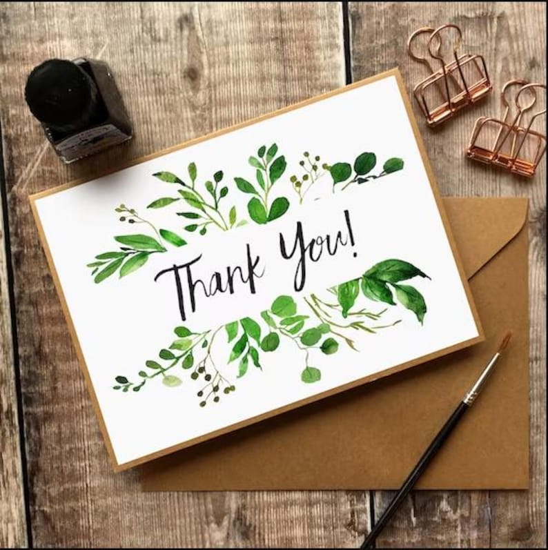 May include: A white card with the words "Thank You!" in black script, surrounded by green watercolor leaves. Includes a brown envelope, ink bottle, paint brush, and copper binder clips. The card is on a wooden surface.