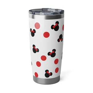 May include: A white travel tumbler with a silver lid and base. The tumbler is decorated with a pattern of black Minnie Mouse bows with red bows and red polka dots. The tumbler is designed to keep drinks hot or cold.