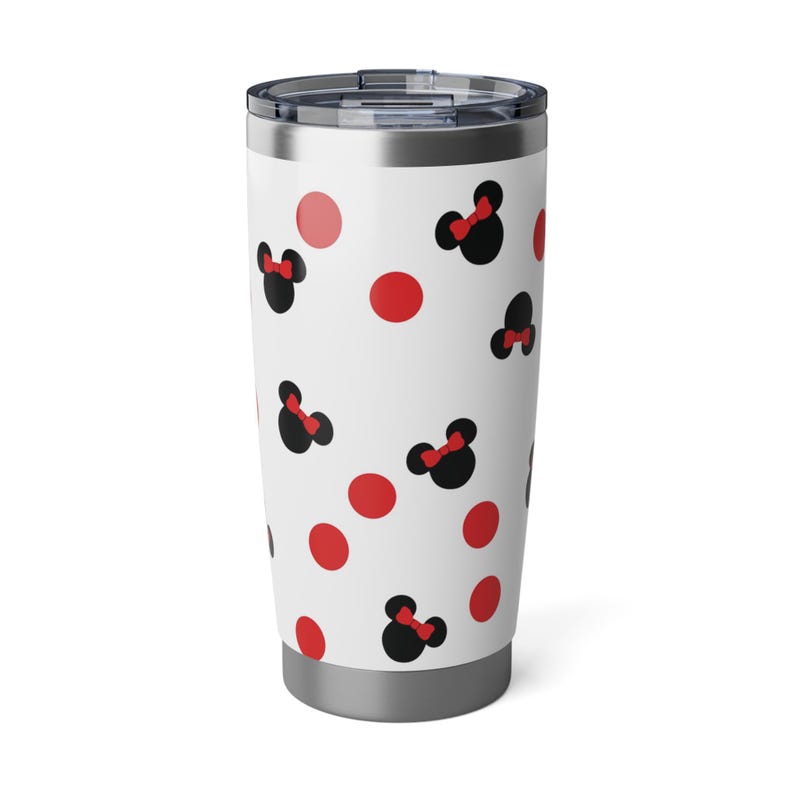 May include: A white travel tumbler with a silver lid and base. The tumbler is decorated with a pattern of black Minnie Mouse bows with red bows and red polka dots. The tumbler is designed to keep drinks hot or cold.