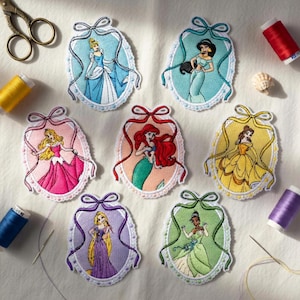 May include: Embroidered patches featuring various princesses in oval frames with lace trim and ribbon bows. Each patch showcases a different princess in vibrant colors, including Cinderella, Jasmine, Aurora, Ariel, Belle, Rapunzel, and Tiana. Sewing supplies are in the background.