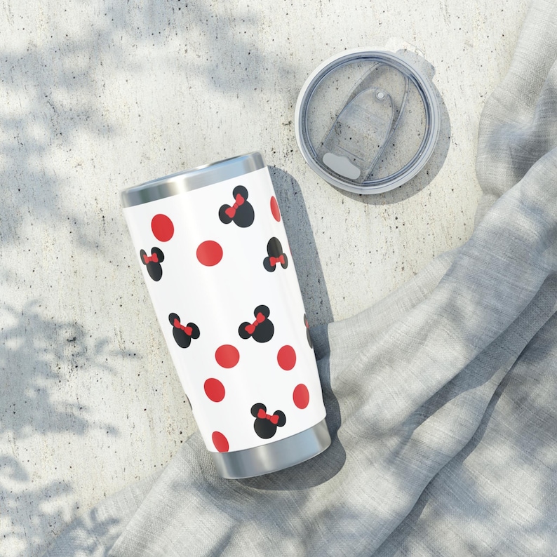 May include: A white tumbler with a silver rim and base, featuring a pattern of red polka dots and black Minnie Mouse bows with red bows. The lid is clear and sits next to the tumbler.
