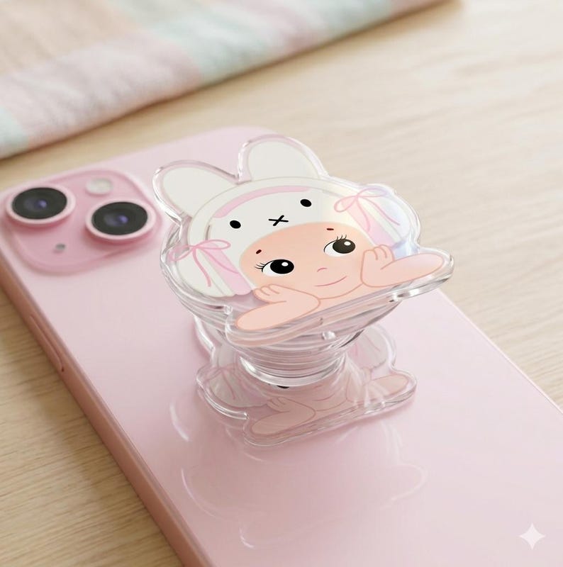 May include: A pink smartphone with a clear phone grip featuring a cartoon bunny design. The bunny has a white hat with pink accents and is resting its chin on its hands. The phone has a dual-lens camera.