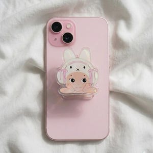 May include: A pink smartphone with a clear, cartoon-style phone grip featuring a baby wearing a bunny hat. The phone grip has a holographic effect. The phone is resting on a white, textured surface.