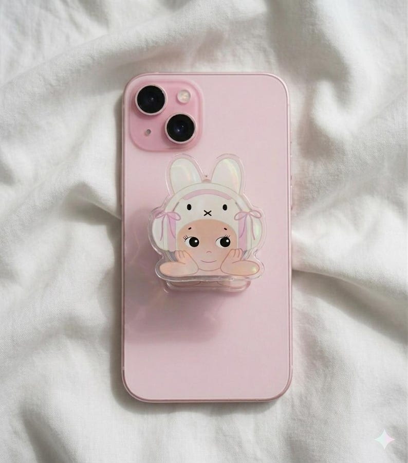 May include: A pink smartphone with a clear, cartoon-style phone grip featuring a baby wearing a bunny hat. The phone grip has a holographic effect. The phone is resting on a white, textured surface.