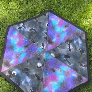 May include: A hexagonal fabric mat with a black border. The mat is divided into six triangular sections. Each section features a different design, including a night sky with dragons, moons, and stars in shades of purple, blue, and gray.