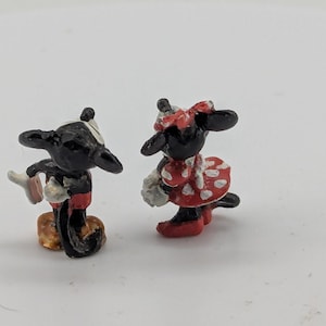 May include: Miniature figurines of Mickey Mouse and Minnie Mouse. Mickey is in black, red, and white, holding a glove. Minnie is in a red dress with white polka dots and a matching bow. Both have black ears.