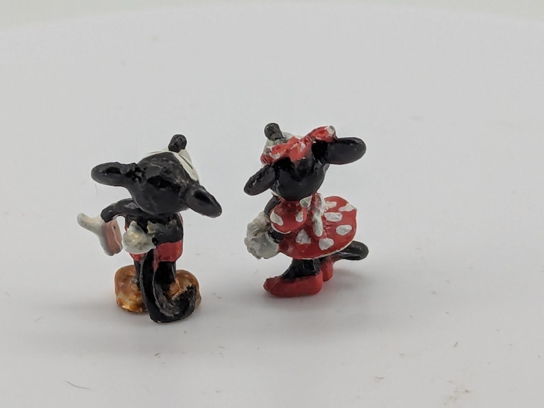 May include: Miniature figurines of Mickey Mouse and Minnie Mouse. Mickey is in black, red, and white, holding a glove. Minnie is in a red dress with white polka dots and a matching bow. Both have black ears.