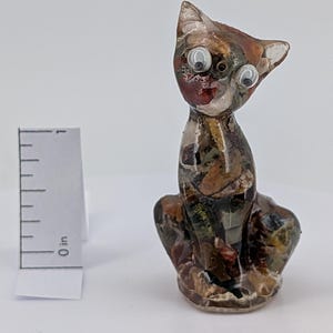 May include: A small, decorative cat figurine with large, googly eyes. The cat is made of a clear material filled with colorful, textured pieces. The figurine is approximately 1 inch tall, sitting upright with its paws tucked in.