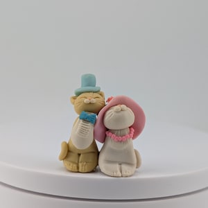 May include: A whimsical figurine set featuring a cat and a dog. The cat is tan with a blue top hat and a blue scarf. The dog is white with a pink hat and a pink beaded necklace. The figurines are on a white surface.