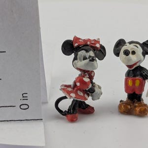 May include: Two miniature figurines of Mickey and Minnie Mouse. Mickey is wearing red shorts and brown shoes. Minnie is wearing a red polka dot dress and bow. Both figures have black ears and are standing on a white surface.