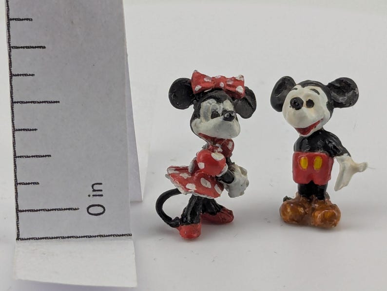 May include: Two miniature figurines of Mickey and Minnie Mouse. Mickey is wearing red shorts and brown shoes. Minnie is wearing a red polka dot dress and bow. Both figures have black ears and are standing on a white surface.