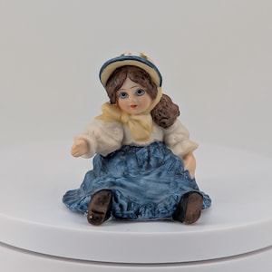 May include: A ceramic figurine of a seated girl wearing a blue and white dress, a yellow scarf, and a hat with floral accents. The doll has brown hair and is posed on a white surface.