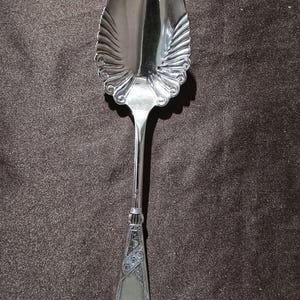 Op de afbeelding: Een zilveren serveerlepel met een decoratieve, geschulpte kom en een sierlijk handvat. De kom van de lepel heeft een gegroefd ontwerp en het handvat is voorzien van gegraveerde patronen. De lepel is ontworpen om te serveren en voegt een vleugje elegantie toe aan elke tafel.