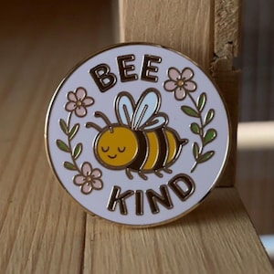 Peut inclure: Épingle ronde en émail avec une bordure dorée. L'épingle représente une abeille de dessin animé avec un corps jaune et des rayures noires, entourée de fleurs roses et de feuilles vertes. Les mots "BEE KIND" sont écrits en or.