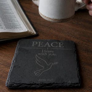 Op de afbeelding: Een vierkante zwarte leistenen onderzetter met een gravure van de tekst PEACE, I leave with you en JOHN 14:27. De onderzetter toont een lijntekening van een duif met een olijftak en heeft ruwe, natuurlijke randen aan alle vier de zijden.