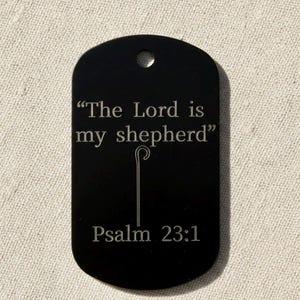 May include: Black dog tag with the engraved text "The Lord is my shepherd" and "Psalm 23:1". A shepherd's crook is etched below the quote. The tag has a rounded rectangular shape with a hole at the top.