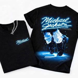 May include: Two black t-shirts. One shirt has the name "Michael" in a blue neon font. The other shirt has the name "Michael Jackson" in a blue neon font, with an image of a dancer and the text "DISCOVER THE KING OF POP". A silver chain necklace is on the first shirt.