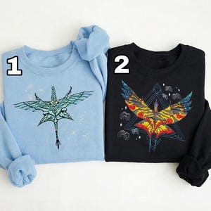 May include: Two sweatshirts, one light blue and one black, each featuring a detailed graphic of a winged creature. The light blue sweatshirt has a teal and black design, while the black one has a vibrant red, yellow, and blue design.
