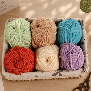 Op de afbeelding: Een rechthoekige, met stof beklede mand met zes bollen zacht fluweelgaren in lichtgroen, beige, felblauw, rood, crème en paars. De mand heeft een witte voering met een subtiele bruine botanische print en een geweven onderkant.