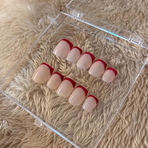 May include: A set of artificial nails in a clear plastic case. The nails are a light pink with a vibrant red French tip design. The nails are arranged in two rows, showcasing the different sizes.