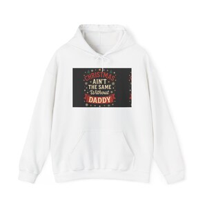 Puede incluir: Sudadera con capucha blanca con bolsillo delantero. La sudadera presenta un gráfico con el texto "Christmas ain't the same without DADDY" en rojo, verde y blanco. El gráfico incluye copos de nieve y diseños de árboles de Navidad.